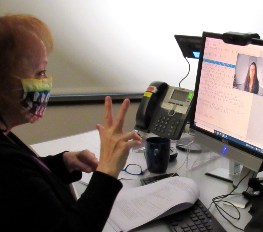 A woman with a COVID-19 mask signing to a computer screen. On the screen is a video call and braille language