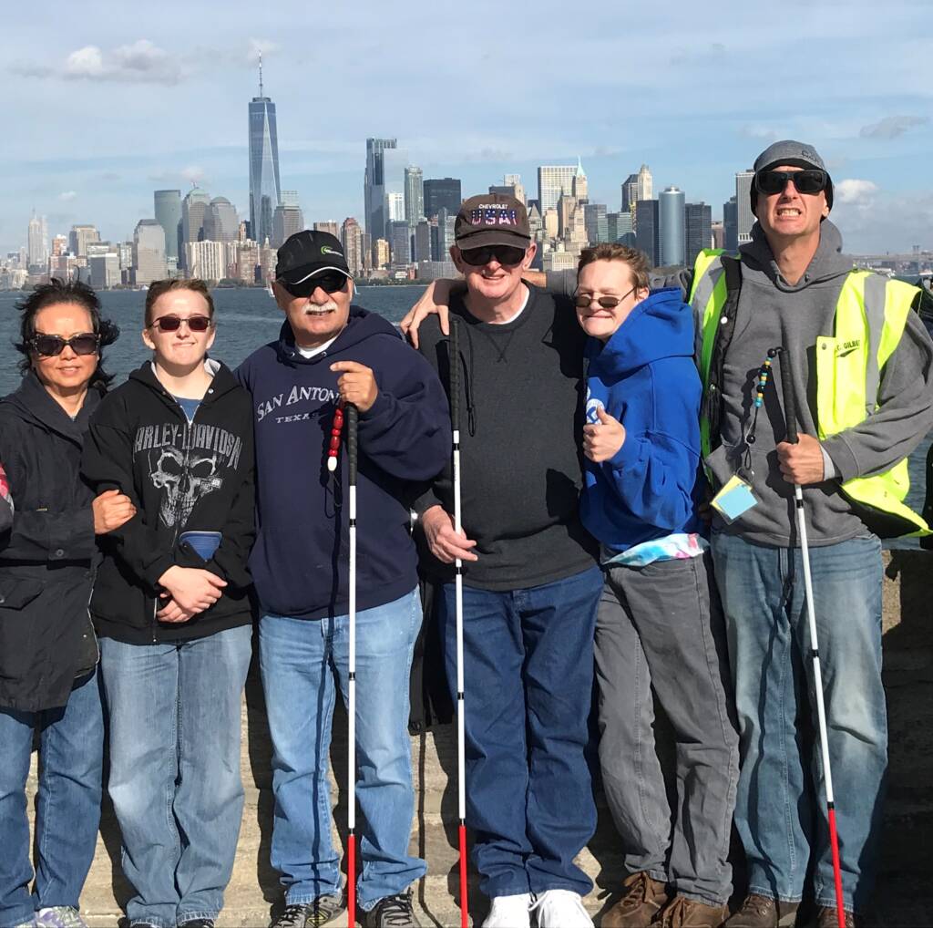 Six people wearing sunglasses and smiling in front of a city skyline. Three of the people are holding white canes