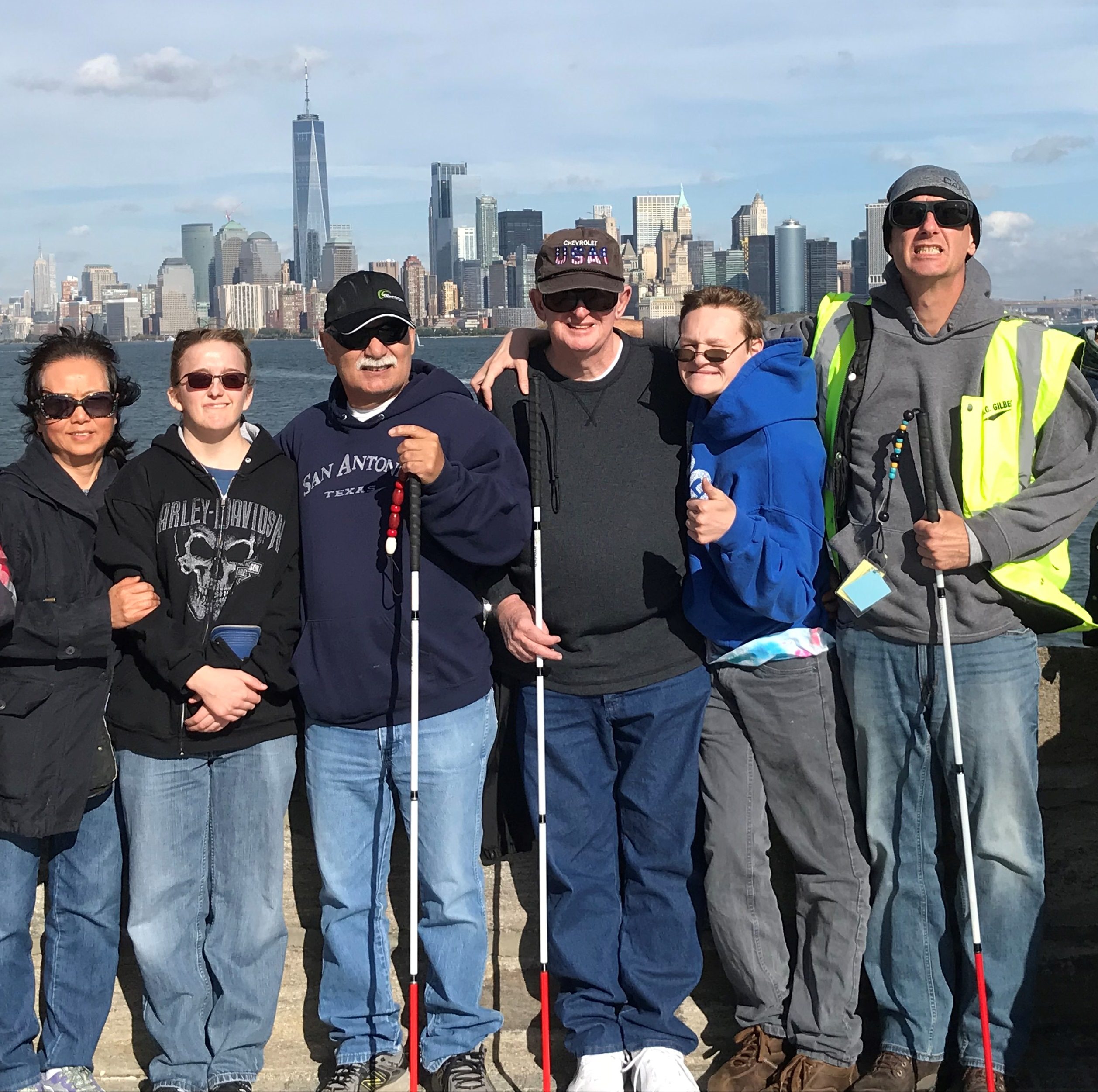 A group of men and women, some holding white canes, stand in front of the NY City skyline.