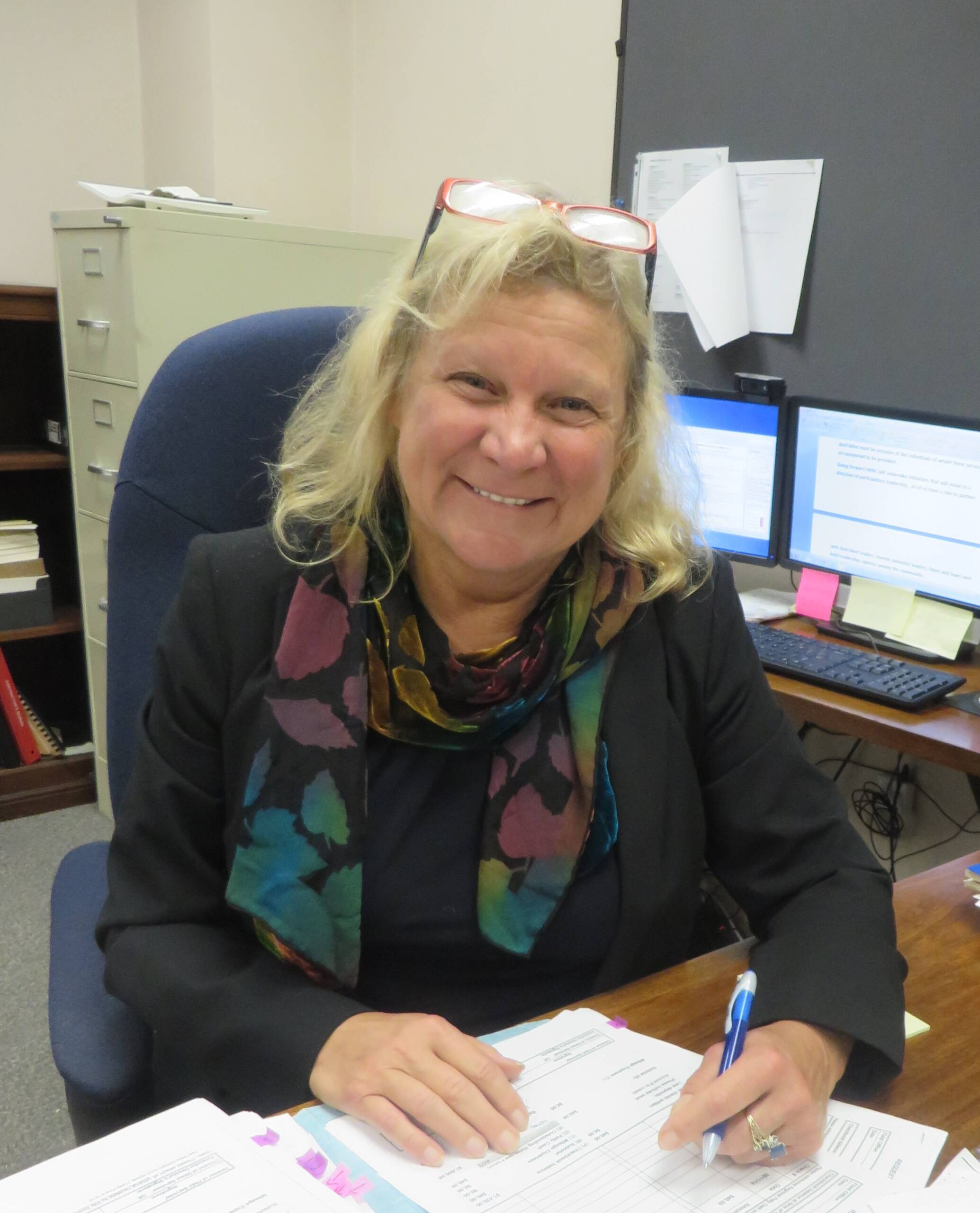 A woman with blond hair smiling and wearing a black jacket sits at a desk.