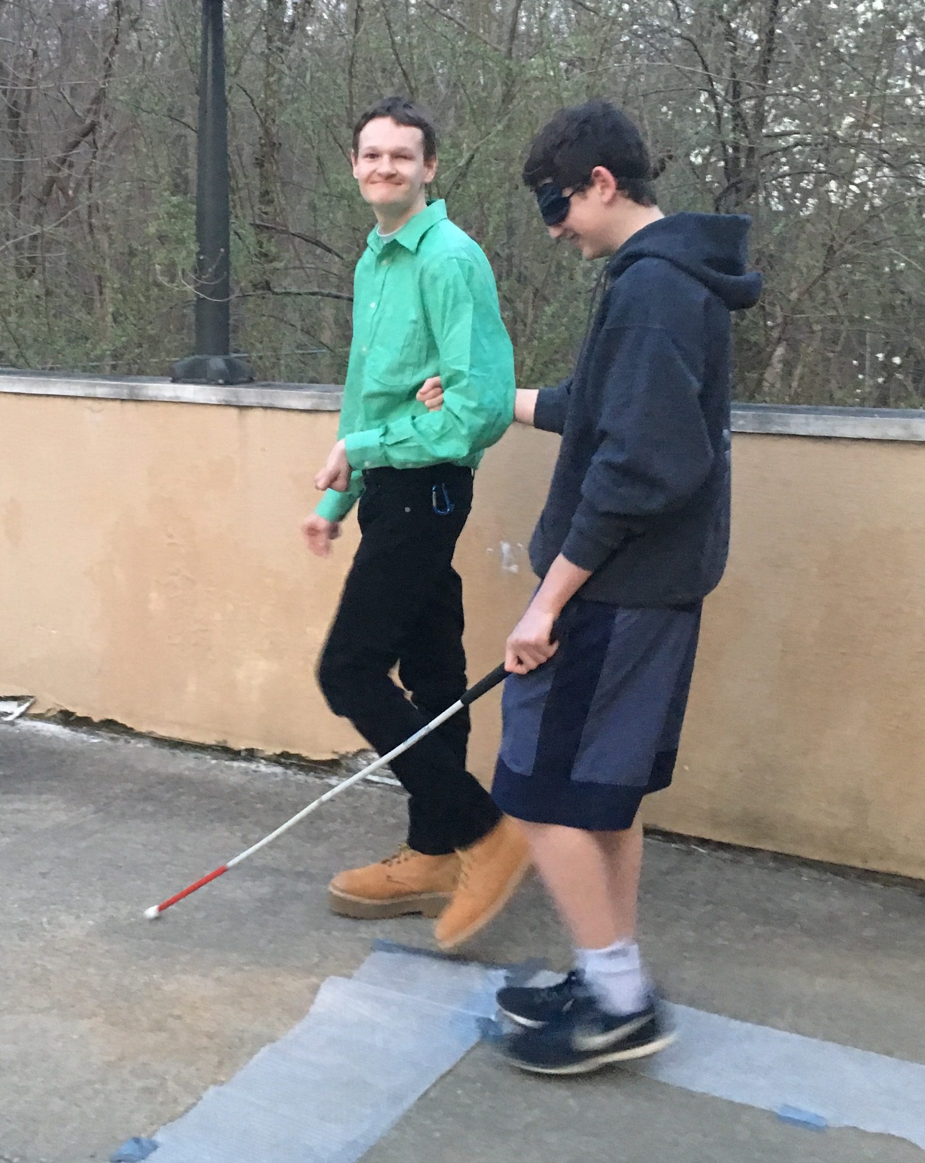 A young man guides another young man who is wearing a blindfold and holding a white cane.