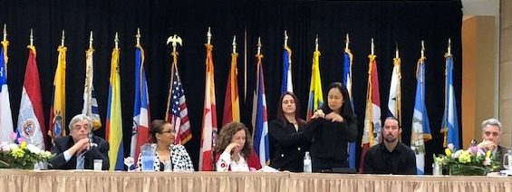 Two women stand at a large table with men and women sitting on either side of them. Behind them are several colorful flags on stands.