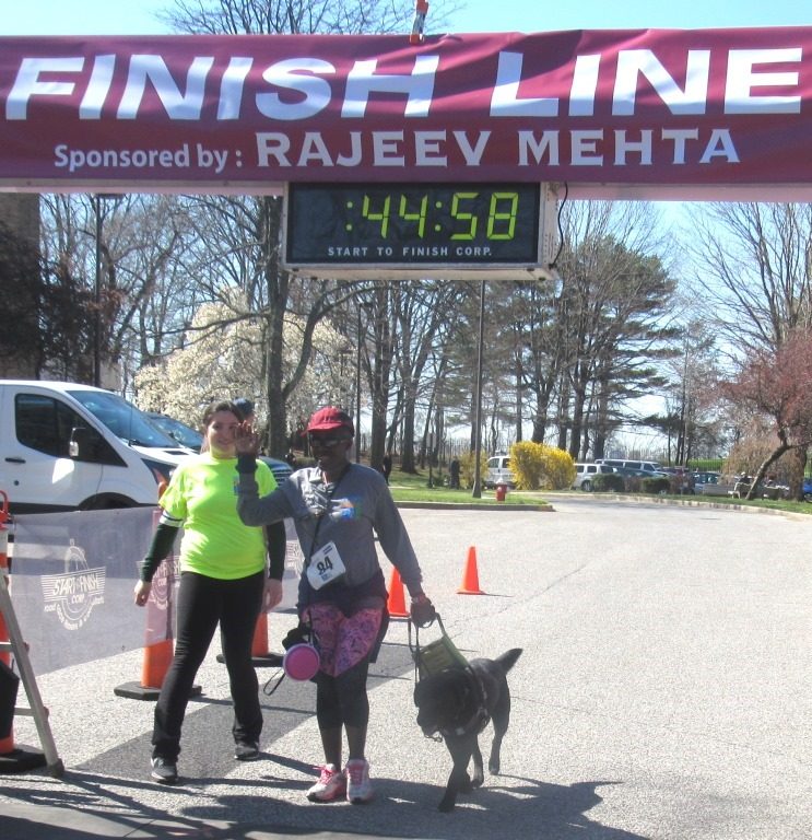 A woman in a red cap walking with a dog guide and a woman in a yellow t-shirt cross the finish line.