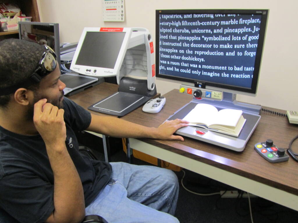 A man reading a book using a closed caption tv that enlarges the print onto a screen