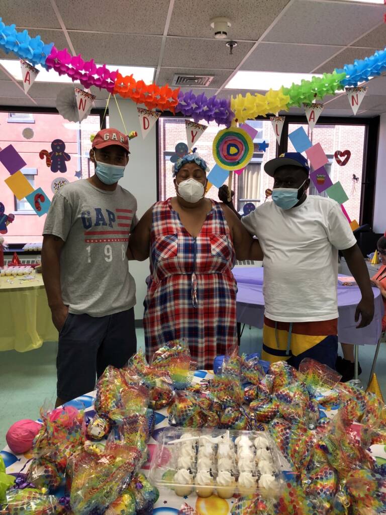 3 people standing in room with arms around each other surrounded by rainbow decorations. On the windows behind them are gingerbread men, hearts, candy canes, and colorful rectangles. In the foreground in front of the 3 people is a table with a balloon-patterned table cloth and treats on top of it