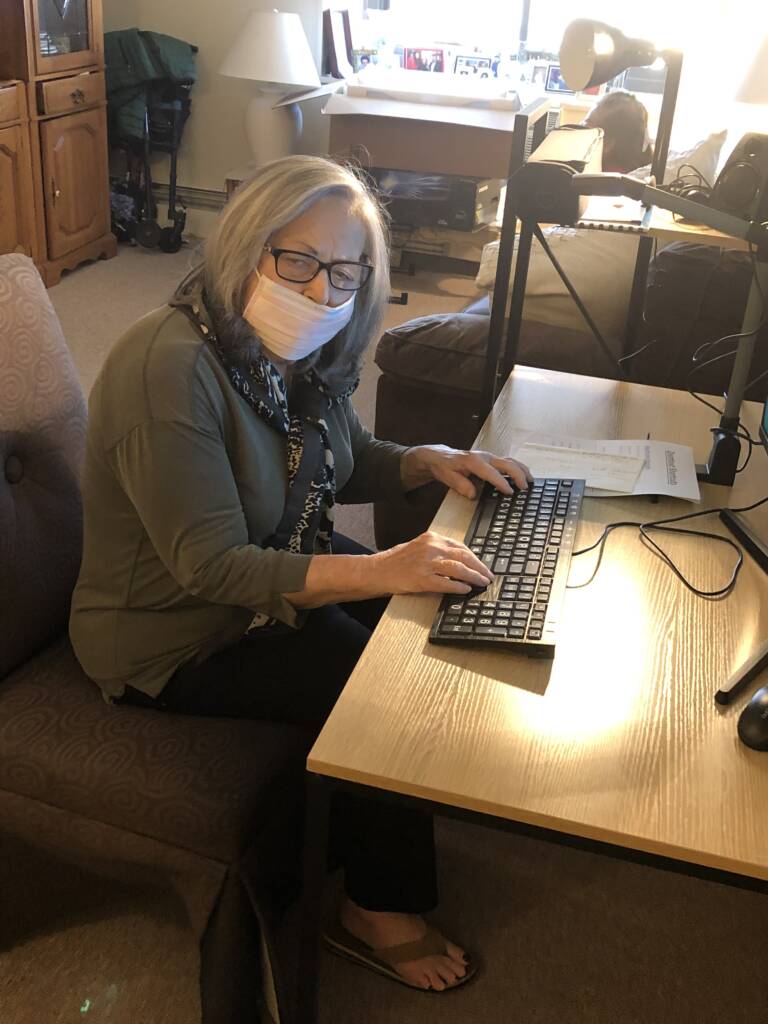 An older woman named Barbara Tucker wearing a facemask and sitting at a desk with her hands on a computer keyboard