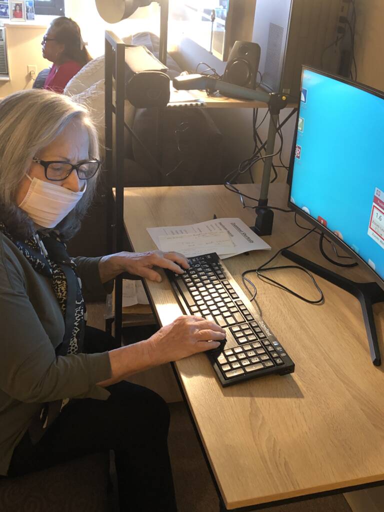 An older woman named Barbara Tucker wearing a facemask and sitting at a desk with her hands on a computer keyboard
