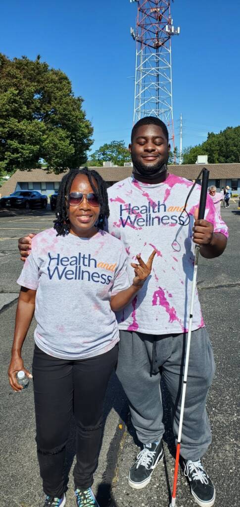 A woman wearing sunglasses and a man holding a white cane smiling outside wearing shirts that say Health and Wellness on them