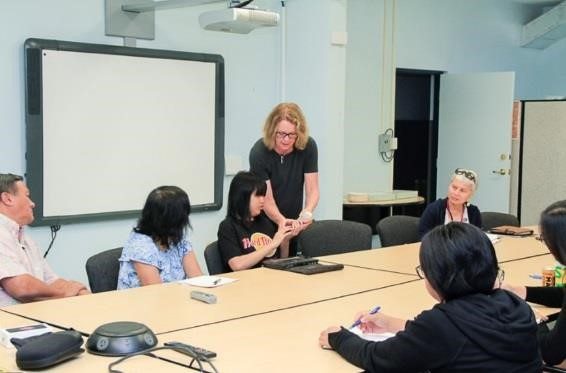 A woman named Cathy standing teaches DeafBlind awareness to agency personnel sitting around a conference table