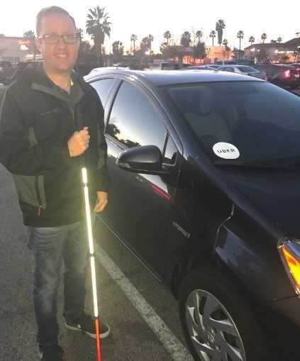 Chad standing in front of an Uber car holding his white cane.
