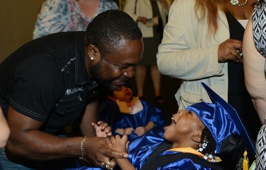 A man smiling and holding a young boy’s wrist. The young boy is also smiling back at the man and is wearing a blue graduation cap and gown