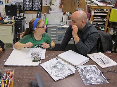 Male instructor and young girl wearing a hearing aid and glasses sitting in art studio surrounded by pencil drawings