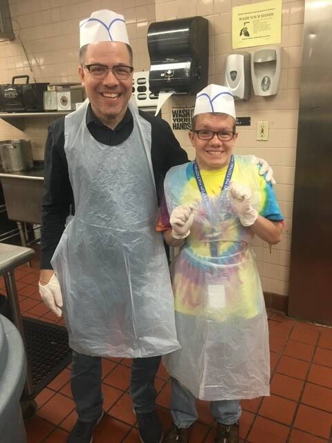 2 people smiling in a restaurant-style kitchen wearing kitchen hats, glasses, and translucent aprons 