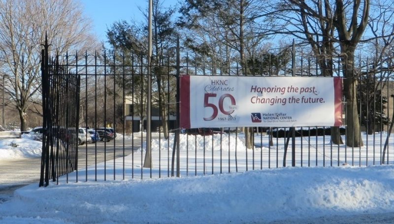 Sign outside that says Group of people standing outside of the Helen Keller National Center holding a large sign that says HKNC Celebrates 50 Years. Honoring the Past, Changing the Future