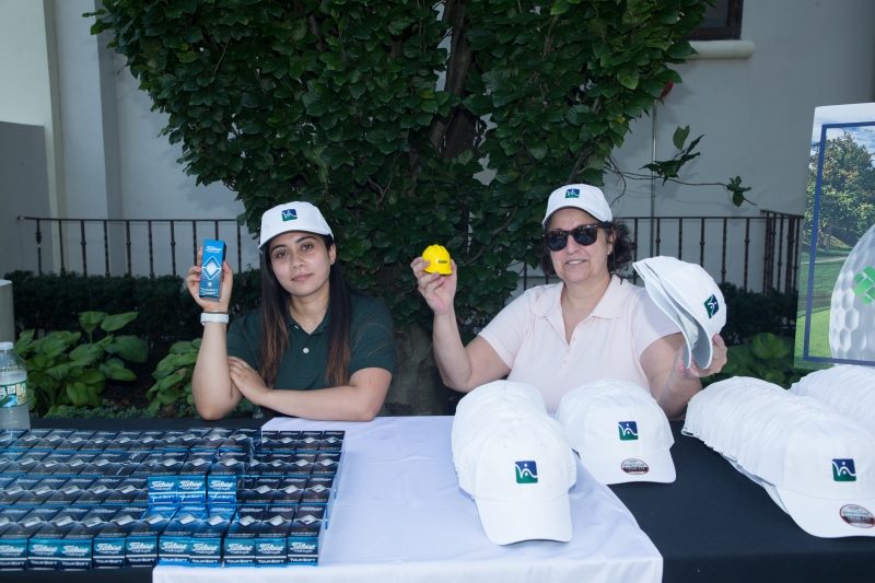A young woman in a HKS white cap is sitting and holding up a sleeve of golf balls with one hand. Another woman is wearing a HKS white cap and holding up a yellow stress ball and two white HKS caps. They are both behind a table with caps and golf ball packages.