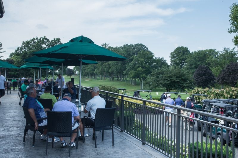 Groups of individuals sitting outdoors under a table with shading. Below the balcony are individuals getting into golf carts.