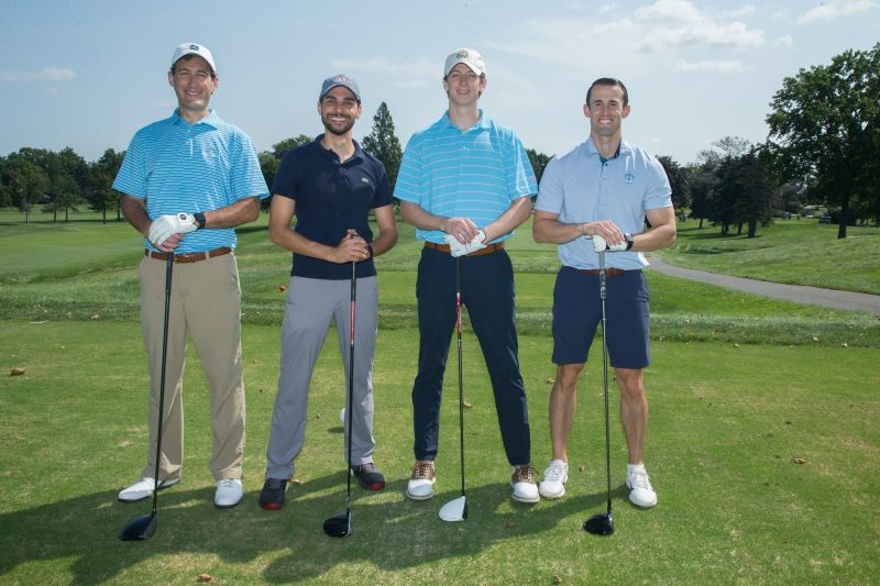 Four men standing on a golf course. Three wear caps, polo shirts, and pants. One wears a polo shirt and shorts. They are all holding onto their golf clubs.