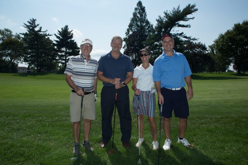 Three men and a woman with their golf clubs on a golf course.