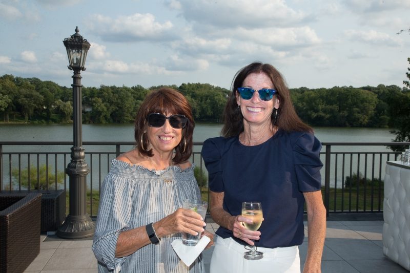 Two woman with a lake view behind them are holding drinks.