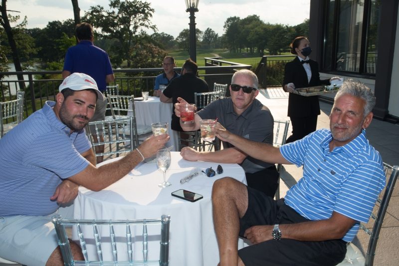 Three men cheering their drinks at a table outdoors.