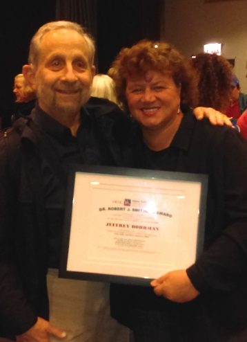 A man and woman stand close to each other while the woman holds a framed award