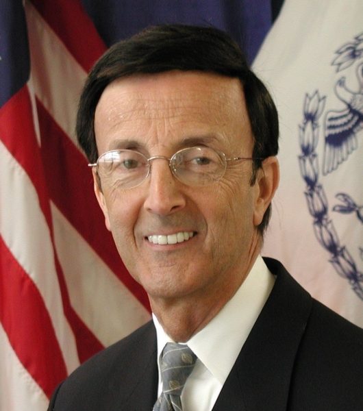 Joseph F. Bruno, a man wearing glasses and a suit and tie, smiles in front of an American flag