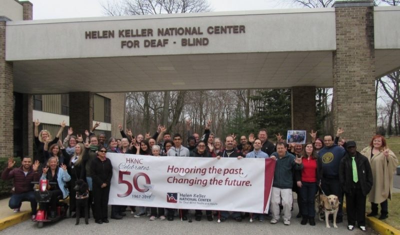 Group of people standing outside of the Helen Keller National Center holding a large sign that says HKNC Celebrates 50 Years. Honoring the Past, Changing the Future