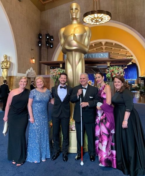 A group of two men and 4 women with light skin stand in front of a statue of Oscar which is twice the size of the people. They are all wearing formal wear - the men in tuxedos with bow ties and the women in floor length gowns. One man is holding a white cane