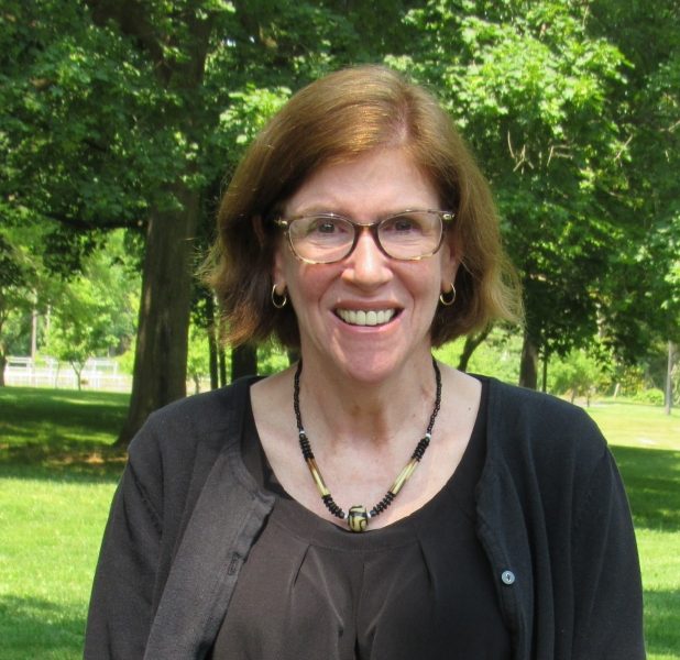 Head shot of a woman with light skin, shoulder length straight red hair wearing glasses, a black blouse and black and white necklace with green trees in the background
