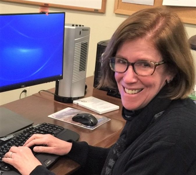 Head shot of a woman with light skin, shoulder length straight red hair wearing glasses and a black blouse. She is sitting at a computer with her hands on the keyboard