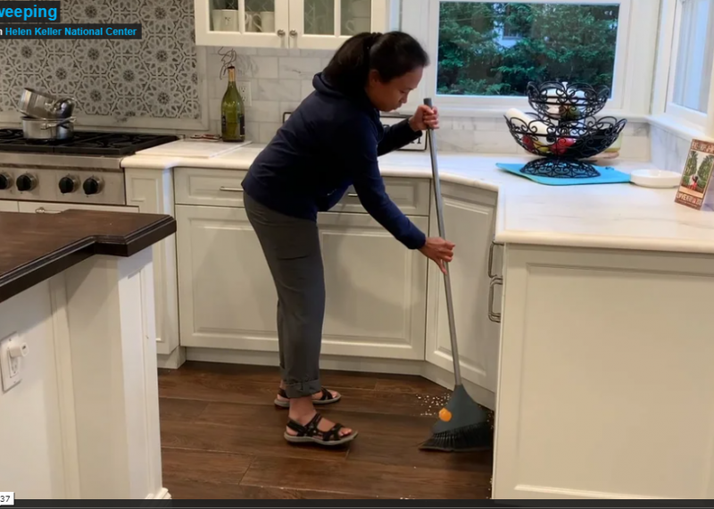 A woman with black shoulder length hair wearing a dark shirt and pants is holding a broom in the corner of a kitchen with white cabinets