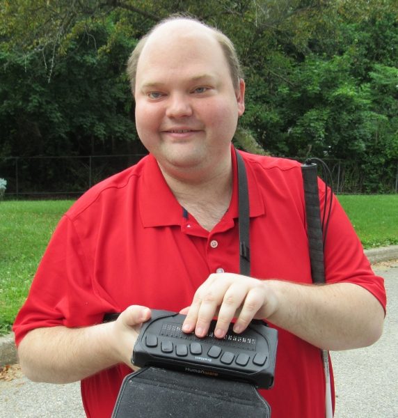 Head shot of a man with light skin and receding short brown hair wearing a red collared t-shirt. He is holding a refreshable braille display with one hand and the other hand on the braille keys