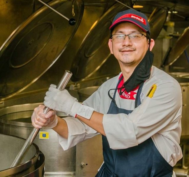 A man with light skin wearing a blue and red Sodexo hat and a white shirt with an apron over it. He has his two hands on a large metal rod suspended into a large pot