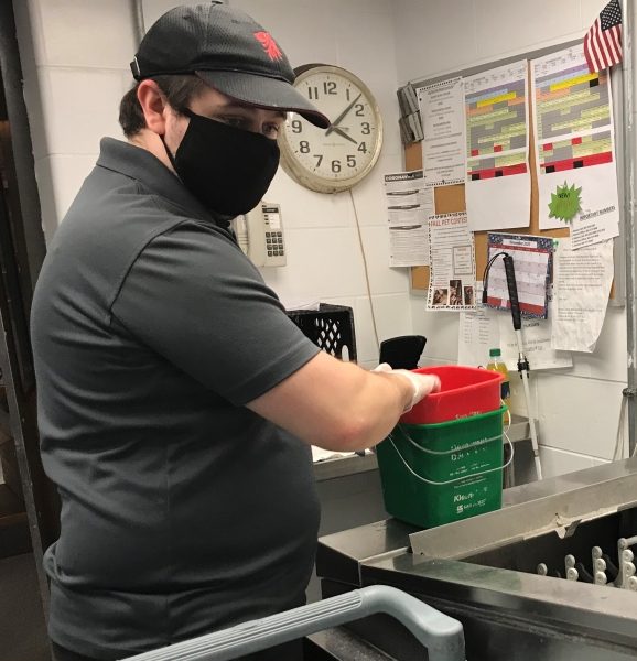 A man with light skin wearing a baseball cap, black face mask and black t-shirt is standing in a kitchen with his hand in a container