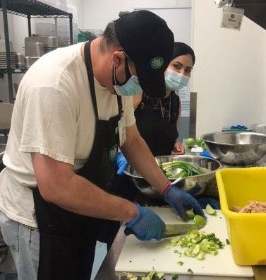 A man with light skin stands at a counter chopping a green vegetable. He is wearing a white t-shirt with a black apron, a black baseball hat and facemask. A woman in black wearing a facemask stands next to him.
