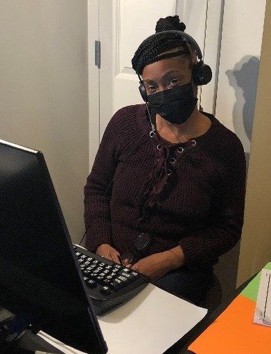 A woman with dark skin dressed in black wearing a black face mask sits at a computer