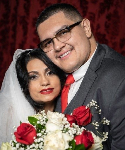 Head shot of a woman with light skin in a bridal veil with long black hair and red lipstick hugging a man with light skin, short black hair wearing a suit jacket and red tie. They are holding a bouquet of red and white roses
