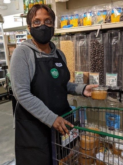 A woman with a grey shirt and a black apron stands with a shopping cart. She is wearing a black face mask.
