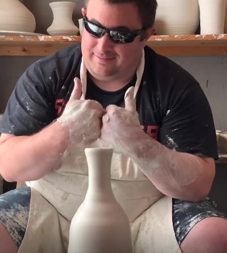 A young man with light skin and short brown hair wearing sunglasses, a black t-shirt and white apron sits behind a clay vase with his clay covered hands giving the thumbs up