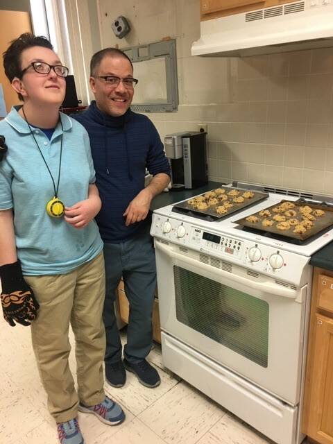 Two people standing in kitchen in front of cookies that are on stovetop; the young person has a kitchen timer around their neck and the man has his arm around the young person