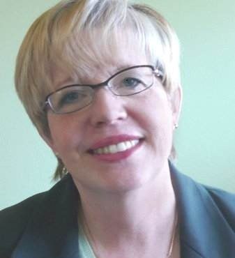 Kathy West-Evans smiling at the camera. She has short blonde hair, glasses, and is wearing a suit jacket.
