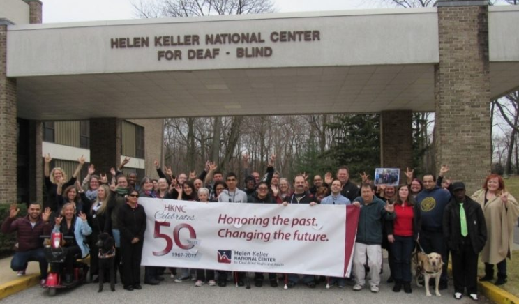 Group of people standing outside of the Helen Keller National Center holding a large sign that says HKNC Celebrates 50 Years. Honoring the Past, Changing the Future