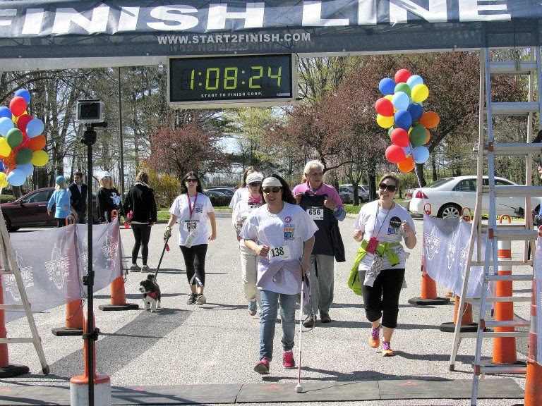 Adults walking outside and crossing the finish line at Helen's 5K Run/Walk event. The woman closest to the finish line holds a white cane.
