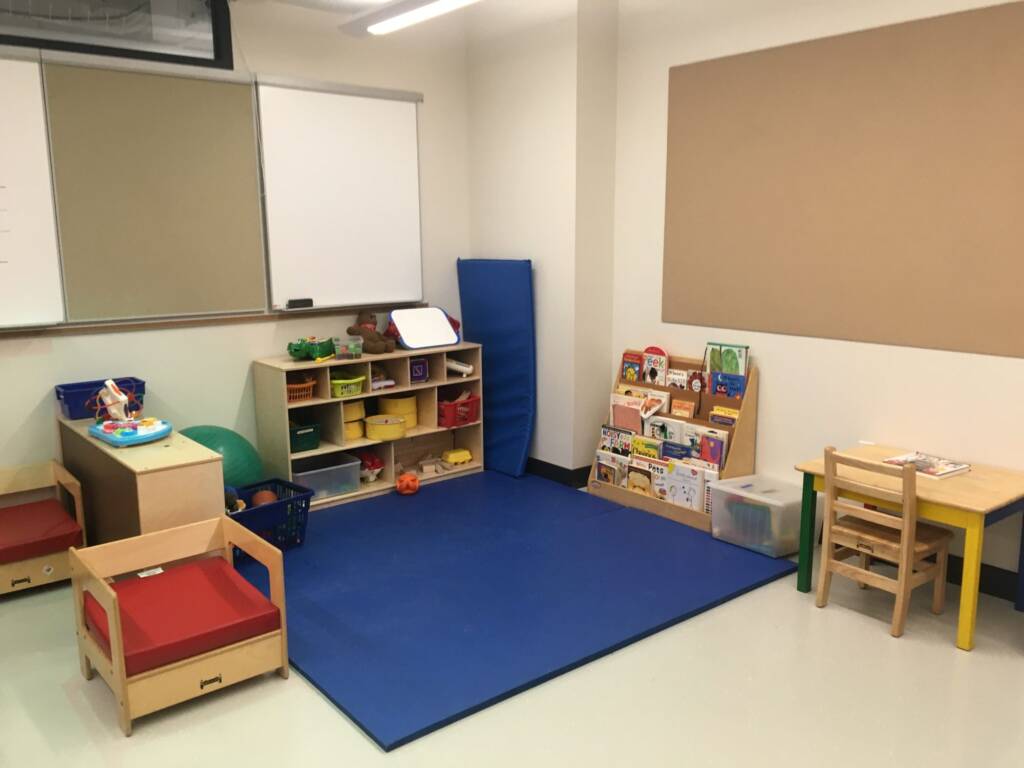 A preschool classroom showing blue mats on the ground and propped against a wall, items in a shelf, a bookcase, and a small desk and chair. 