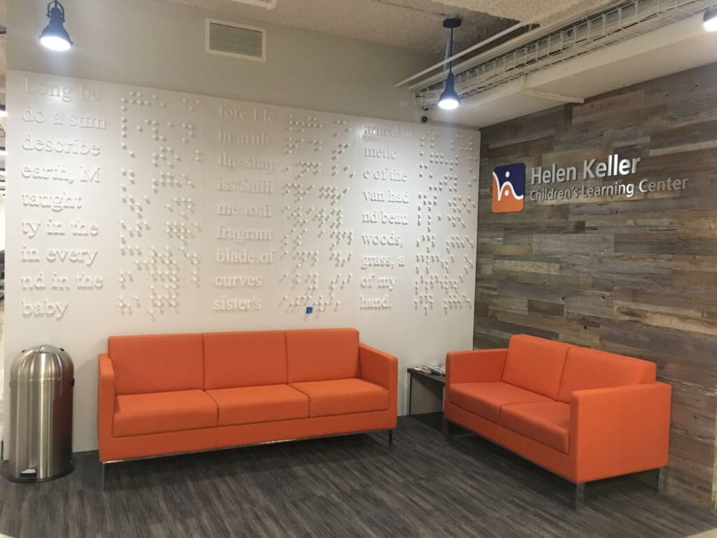 The waiting area of the Children's Learning Center. In the waiting area there are 2 orange couches, a white wall with braille on it to the left, and a Helen Keller Children's Learning Center sign mounted to a wooden wall on the right..