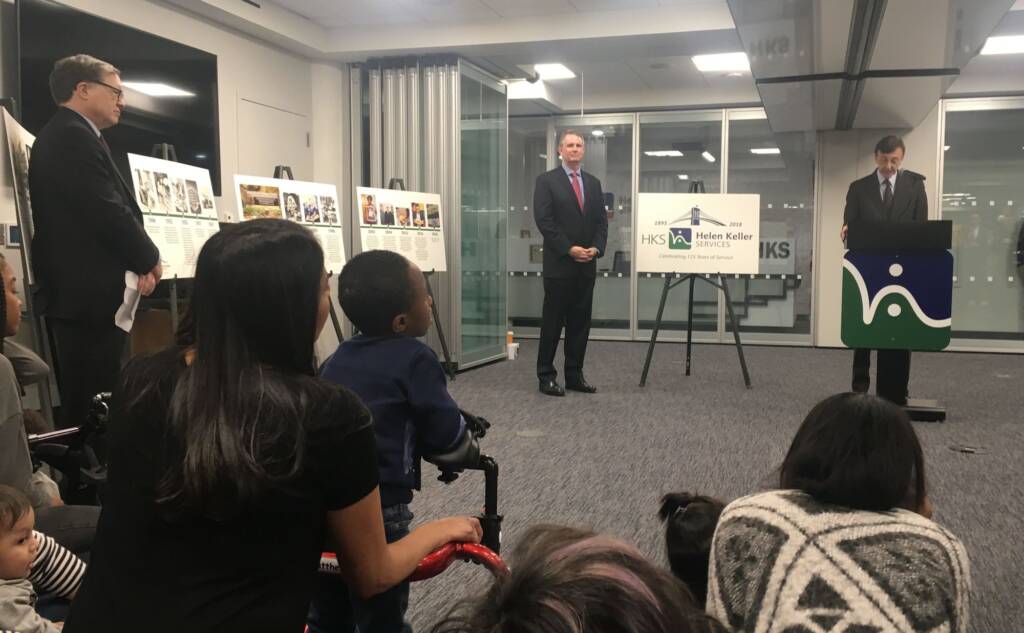 A group of people looking at a man who is speaking behind a podium with the Helen Keller Services logo on it