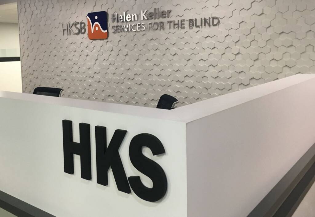 An office space showing a Helen Keller Services for the Blind sign on a textured wall. In front of the wall is a desk with large letters saying HKS on the front of it