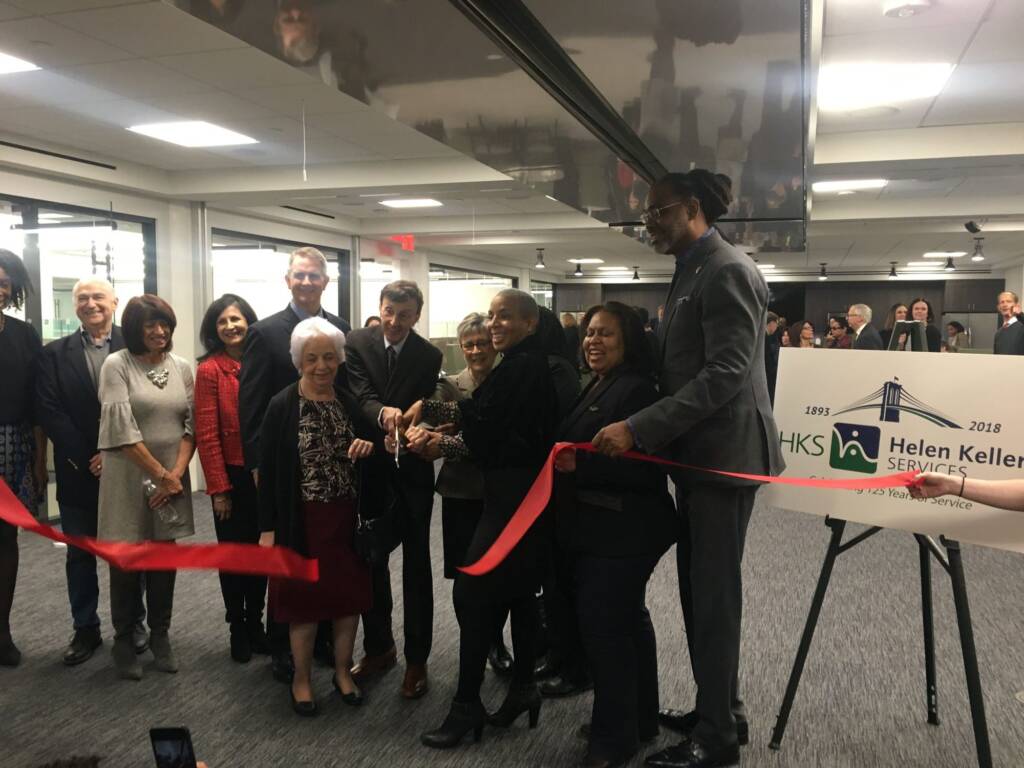 A group of smiling men and women. Many of them hold a scissor together and cut through the middle of a large red horizontal ribbon.