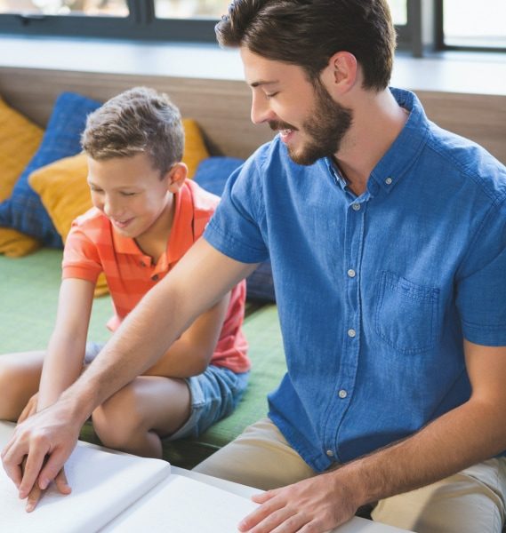 Man and boy sit on a cushion reading a braille book together