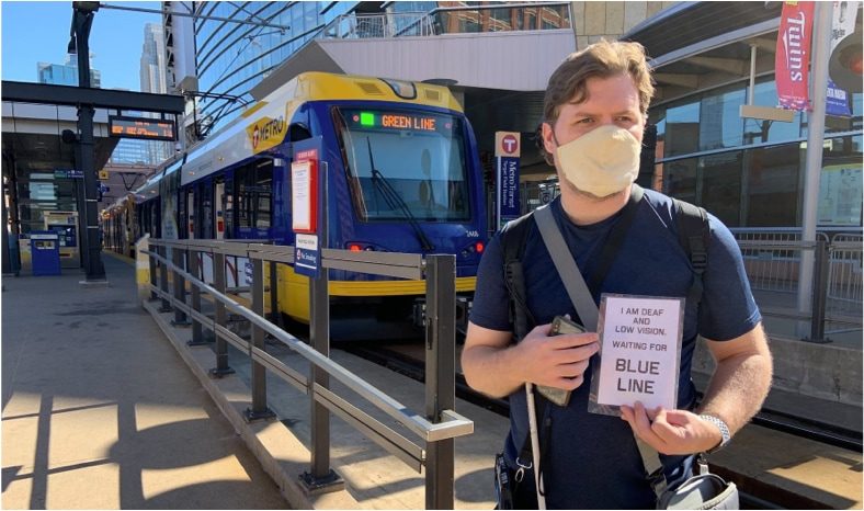 A man who is wearing a COVID-19 mask is standing at a train station, holding a white cane and sign reading: "I am deaf and low vision. Waiting for blue line." He is also wearing a crossbody and holding his cellphone.